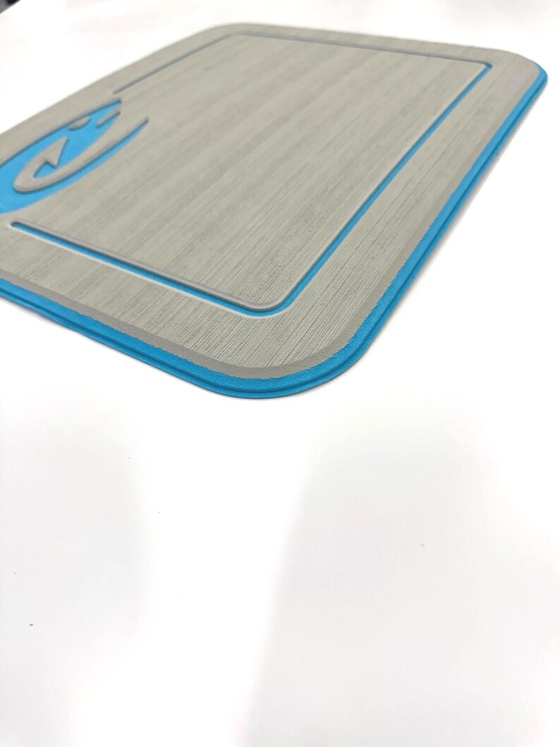 K&ouml;nnte beinhalten: Eine rechteckige graue Matte mit blauem Rand und einem blauen Design in der oberen linken Ecke. Die Matte hat eine strukturierte Oberfl&auml;che und einen vertieften Rand. Das Design scheint ein stilisiertes Wellen- oder Wassersymbol zu sein.