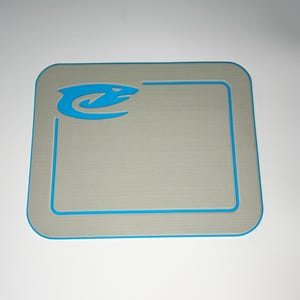 May include: A rectangular, gray mat with a blue border and a blue stylized snake logo in the upper left corner. The mat has a textured surface and rounded corners. The border is a thin, bright blue line.