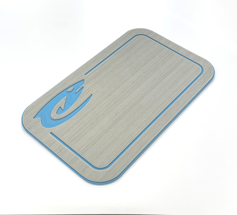 Puede incluir: Tabla de cortar rectangular gris con borde azul y logotipo de pez azul en la esquina superior izquierda. La tabla tiene una superficie texturizada y esquinas redondeadas. Mide aproximadamente 30 cm de largo y 20 cm de ancho.