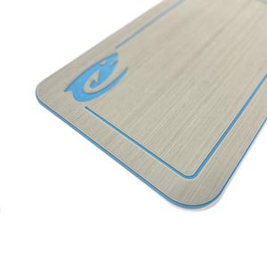 Puede incluir: Una superficie rectangular, de color gris claro texturizado, con un borde azul y un logotipo de pez estilizado azul. La superficie tiene esquinas redondeadas y una sutil apariencia de veta de madera. Los detalles azules contrastan con el fondo neutro.