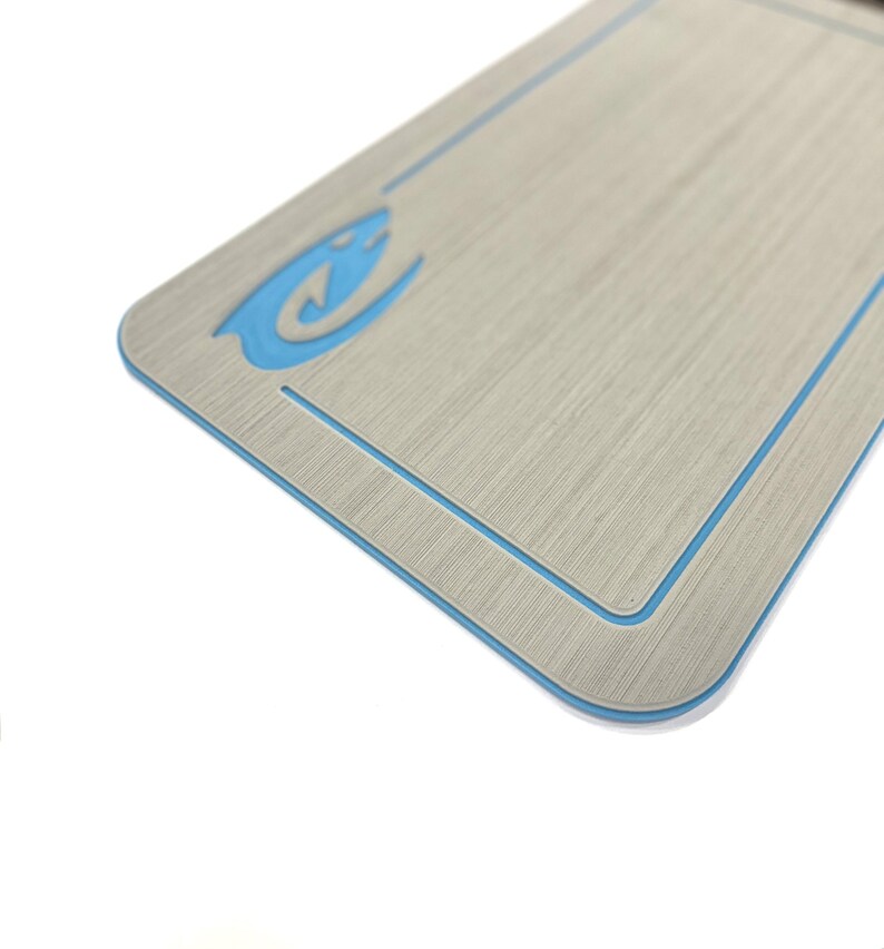 Puede incluir: Una superficie rectangular, de color gris claro texturizado, con un borde azul y un logotipo de pez estilizado azul. La superficie tiene esquinas redondeadas y una sutil apariencia de veta de madera. Los detalles azules contrastan con el fondo neutro.