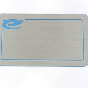 Puede incluir: Una alfombra rectangular gris claro con un borde azul y un logotipo azul estilizado en la esquina superior izquierda. La alfombra tiene esquinas redondeadas y una textura de vetas de madera. El logotipo parece ser un animal estilizado.