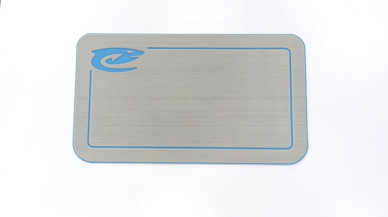 Puede incluir: Una alfombra rectangular gris claro con un borde azul y un logotipo azul estilizado en la esquina superior izquierda. La alfombra tiene esquinas redondeadas y una textura de vetas de madera. El logotipo parece ser un animal estilizado.