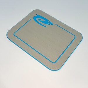 May include: A rectangular, light gray mat with a blue border and a blue stylized design in the upper right corner. The mat has a brushed metal appearance and rounded corners. The blue design is a curved, abstract shape.
