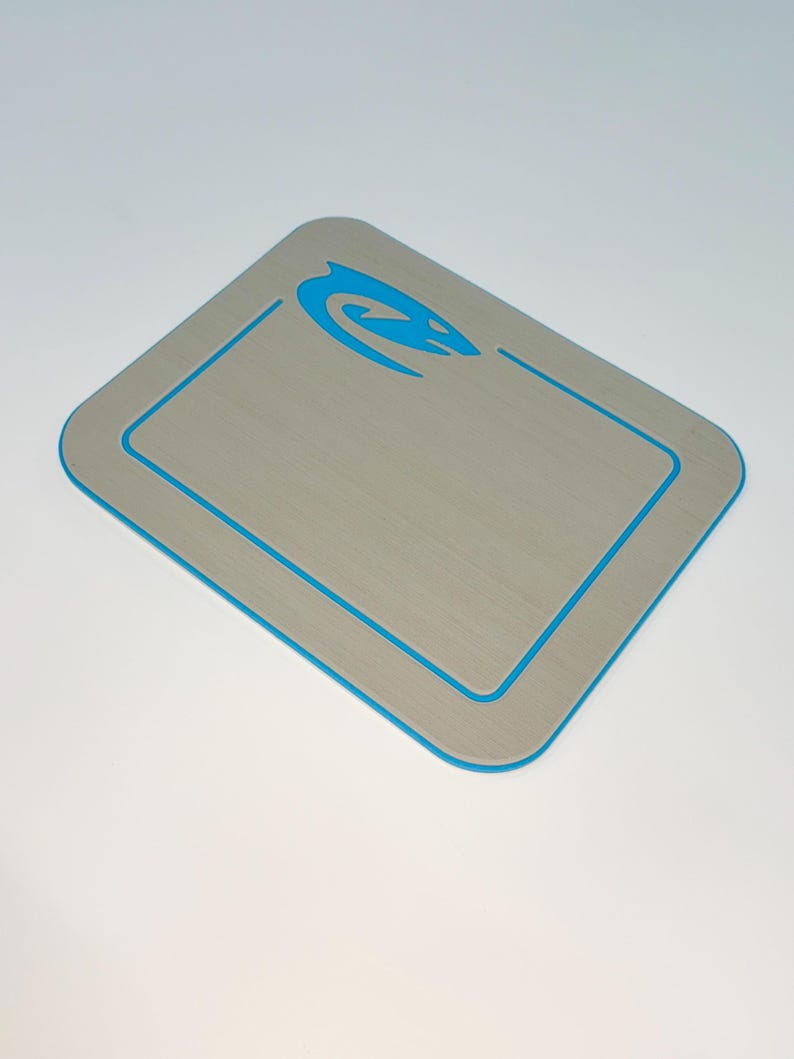 May include: A rectangular, light gray mat with a blue border and a blue stylized design in the upper right corner. The mat has a brushed metal appearance and rounded corners. The blue design is a curved, abstract shape.