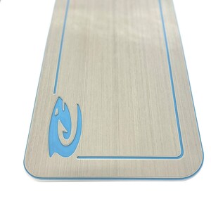 Puede incluir: Una tabla rectangular de madera con un acabado de veta de madera clara. Presenta un borde azul claro y un dise&ntilde;o estilizado de pez u ola azul claro en la esquina inferior izquierda. La tabla tiene esquinas redondeadas.