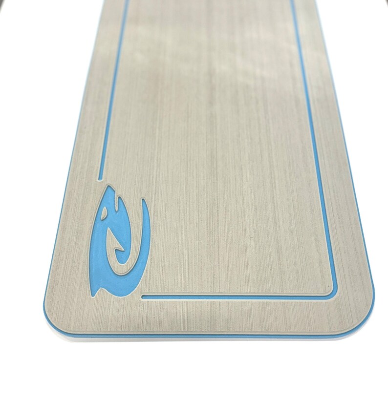 Puede incluir: Una tabla rectangular de madera con un acabado de veta de madera clara. Presenta un borde azul claro y un dise&ntilde;o estilizado de pez u ola azul claro en la esquina inferior izquierda. La tabla tiene esquinas redondeadas.