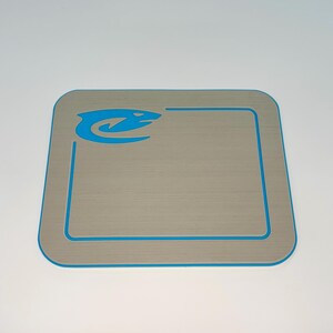 May include: A square, light gray mat with a blue border and a blue logo in the upper left corner. The mat has a textured surface and rounded corners. The logo appears to be a stylized fish or aquatic creature.