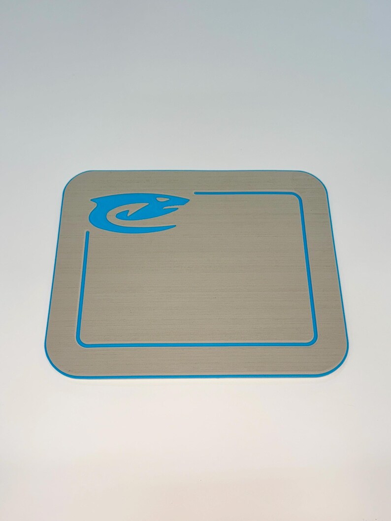 May include: A square, light gray mat with a blue border and a blue logo in the upper left corner. The mat has a textured surface and rounded corners. The logo appears to be a stylized fish or aquatic creature.