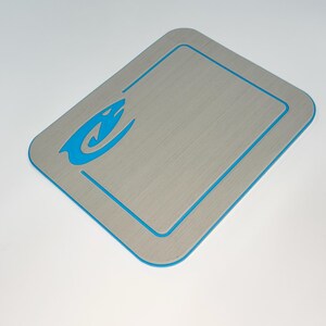 May include: A rectangular, gray mat with a brushed metal appearance. It features a blue border and a blue stylized graphic in the upper left corner. The mat has rounded corners and a clean, modern design.