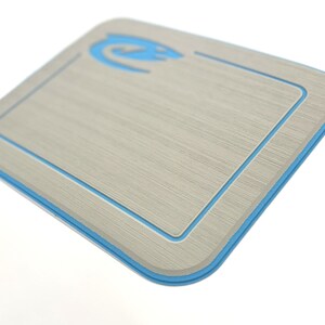 K&ouml;nnte beinhalten: Rechteckiges, hellgraues Schneidebrett mit blauem Rand und blauem Logo in der oberen linken Ecke. Das Brett hat eine strukturierte Oberfl&auml;che und abgerundete Ecken. Das Design ist modern und sauber.