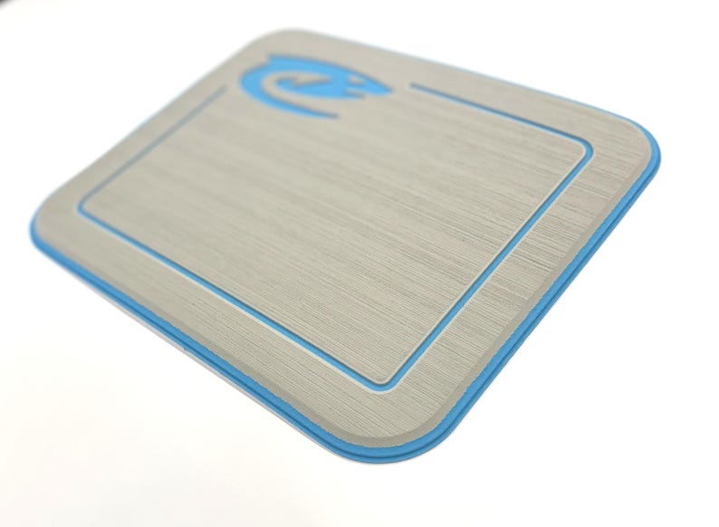 K&ouml;nnte beinhalten: Rechteckiges, hellgraues Schneidebrett mit blauem Rand und blauem Logo in der oberen linken Ecke. Das Brett hat eine strukturierte Oberfl&auml;che und abgerundete Ecken. Das Design ist modern und sauber.