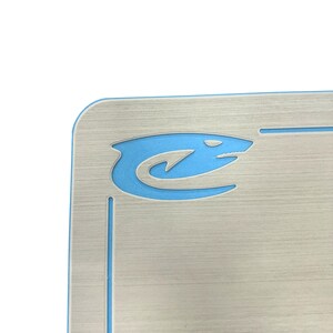 Puede incluir: Placa de matr&iacute;cula plateada con un logotipo de tibur&oacute;n azul en la esquina superior izquierda. El logotipo del tibur&oacute;n es un dise&ntilde;o estilizado con un cuerpo curvo y una cabeza puntiaguda. La placa tiene una textura de metal cepillado.