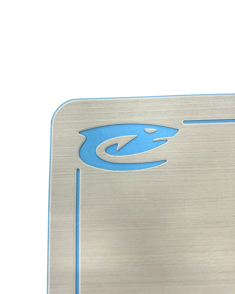 Puede incluir: Placa de matr&iacute;cula plateada con un logotipo de tibur&oacute;n azul en la esquina superior izquierda. El logotipo del tibur&oacute;n es un dise&ntilde;o estilizado con un cuerpo curvo y una cabeza puntiaguda. La placa tiene una textura de metal cepillado.