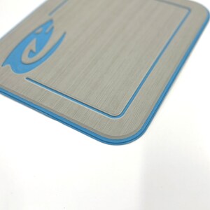 K&ouml;nnte beinhalten: Ein rechteckiges, hellgraues Objekt mit blauem Rand und einem stilisierten blauen Flammendesign in der oberen linken Ecke. Das Objekt hat eine strukturierte Oberfl&auml;che und einen vertieften Innenrand.