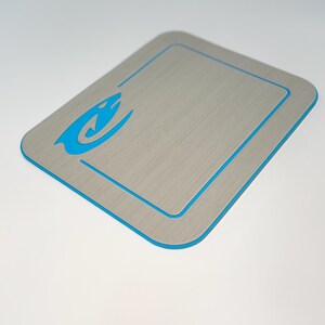 May include: A rectangular, gray cutting board with a blue border and a blue stylized shark logo in the upper left corner. The board has a brushed metal appearance and is designed for kitchen use.