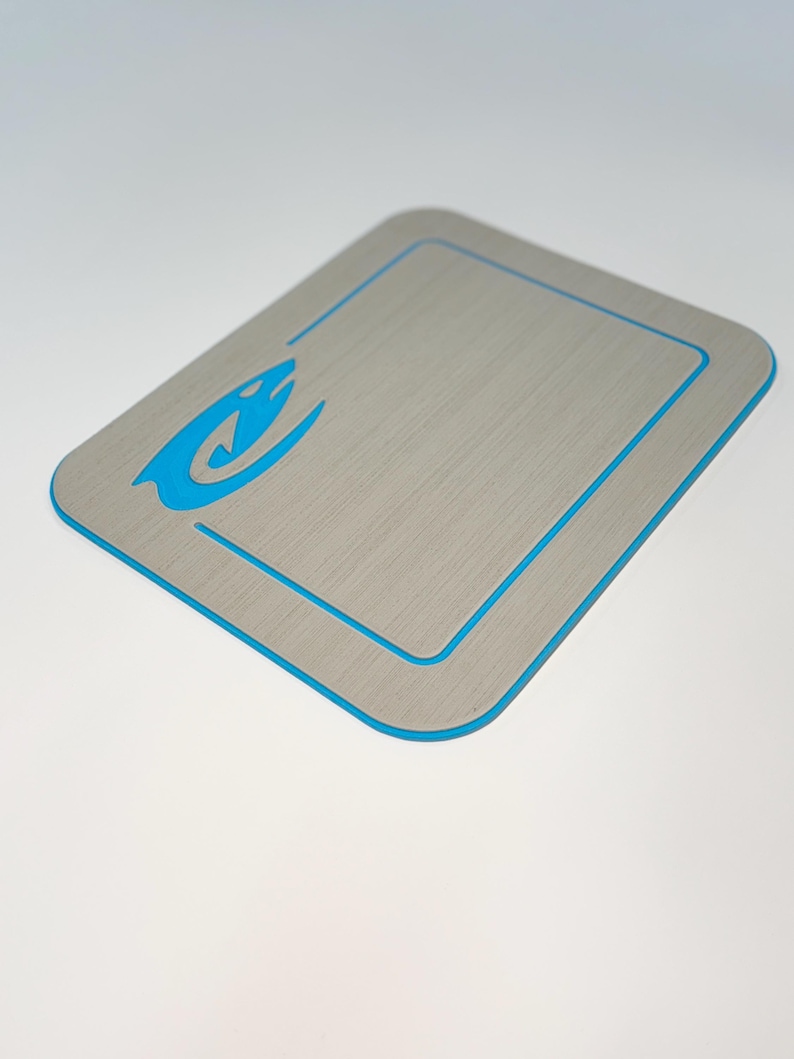May include: A rectangular, gray cutting board with a blue border and a blue stylized shark logo in the upper left corner. The board has a brushed metal appearance and is designed for kitchen use.