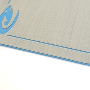 Puede incluir: Una tabla de cortar rectangular con un patr&oacute;n de vetas de madera gris claro y un borde azul. Un gr&aacute;fico azul estilizado est&aacute; en la esquina superior izquierda. La tabla tiene un borde interior empotrado.