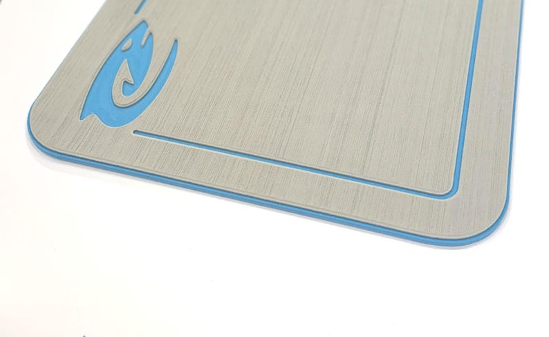 Puede incluir: Una tabla de cortar rectangular con un patr&oacute;n de vetas de madera gris claro y un borde azul. Un gr&aacute;fico azul estilizado est&aacute; en la esquina superior izquierda. La tabla tiene un borde interior empotrado.