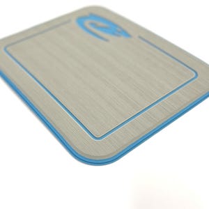 K&ouml;nnte beinhalten: Ein rechteckiges, hellgraues Schneidebrett mit blauem Rand und blauem Logo in der oberen Ecke. Das Brett hat eine Holzmaserung und einen vertieften Innenrand.