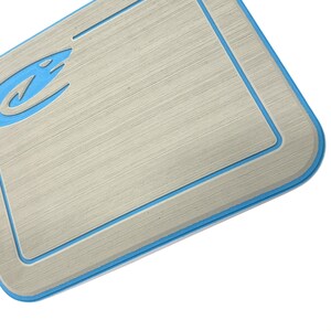 K&ouml;nnte beinhalten: Eine rechteckige, hellgraue Matte mit geb&uuml;rsteter Metalloptik und blauem Rand. Ein blaues Logo befindet sich in der oberen linken Ecke. Die Matte hat abgerundete Ecken und einen vertieften Rand.