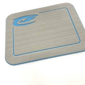 May include: A rectangular, light gray mat with a brushed metal appearance. It features a light blue border and a matching blue logo in the upper left corner. The mat is designed with rounded corners.