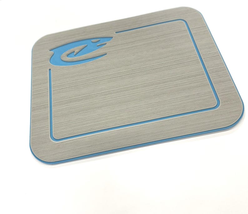 May include: A rectangular, light gray mat with a brushed metal appearance. It features a light blue border and a matching blue logo in the upper left corner. The mat is designed with rounded corners.
