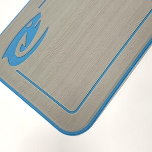 K&ouml;nnte beinhalten: Eine rechteckige, graue Matte mit geb&uuml;rsteter Metallstruktur und blauem Rand. Ein blaues, flammenartiges Design befindet sich in der oberen linken Ecke. Die Matte befindet sich vor einem wei&szlig;en Hintergrund.