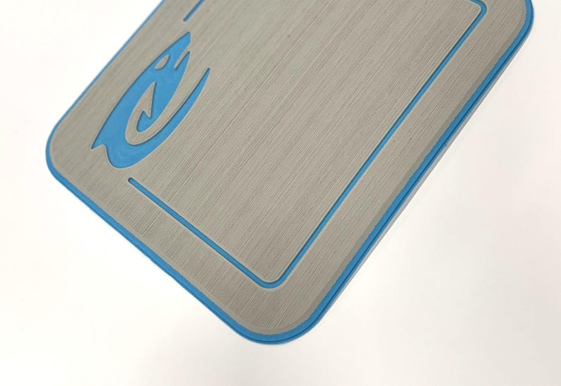 K&ouml;nnte beinhalten: Eine rechteckige, graue Matte mit geb&uuml;rsteter Metallstruktur und blauem Rand. Ein blaues, flammenartiges Design befindet sich in der oberen linken Ecke. Die Matte befindet sich vor einem wei&szlig;en Hintergrund.