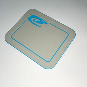 May include: A rectangular, brushed metal-look surface with rounded corners. A thin, bright blue border outlines the edge and a recessed inner rectangle. A blue stylized aquatic creature logo is in the upper right corner.