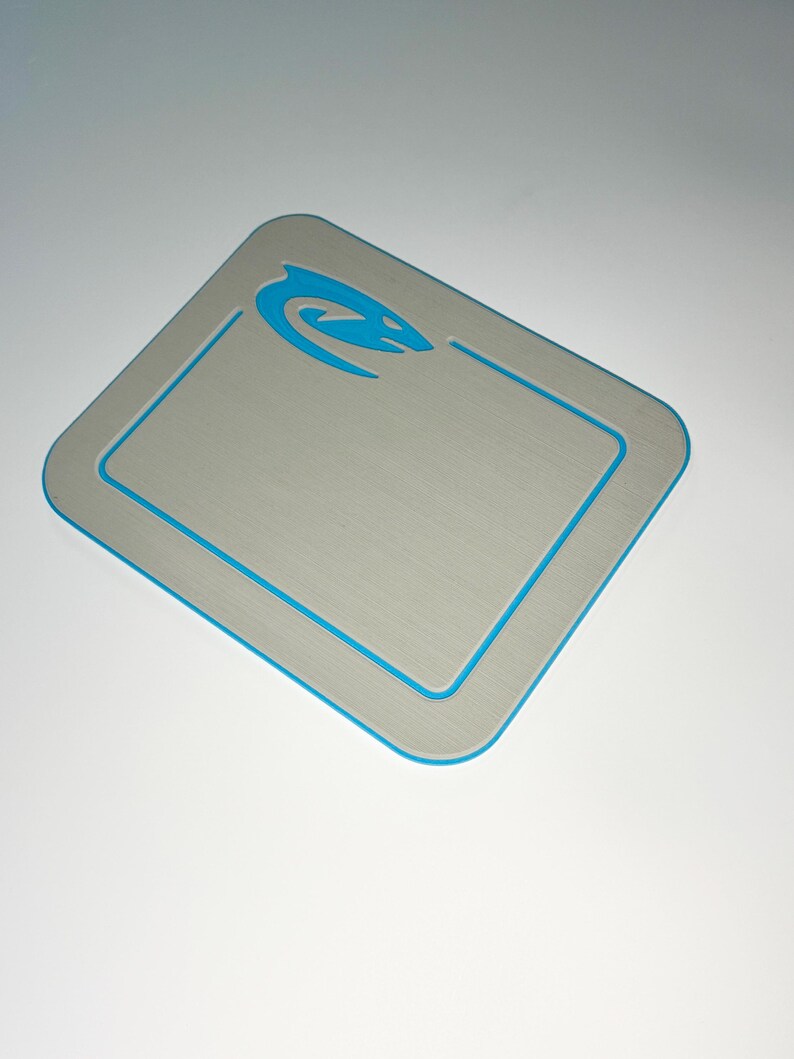 May include: A rectangular, brushed metal-look surface with rounded corners. A thin, bright blue border outlines the edge and a recessed inner rectangle. A blue stylized aquatic creature logo is in the upper right corner.