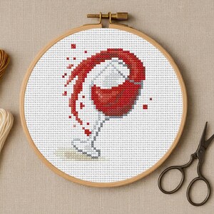 Puede incluir: Un bastidor de bordado con una imagen pixelada de una copa de vino con vino tinto derramándose. Hilos de bordar marrones y crema y unas tijeras pequeñas.