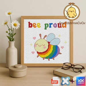 Puede incluir: Una obra de arte de punto de cruz enmarcada con una abeja sonriente de colores del arcoíris y las palabras "bee proud" en letras coloridas. La obra de arte está en un marco de madera, con un jarrón de margaritas y un pequeño recipiente sobre una superficie de madera.