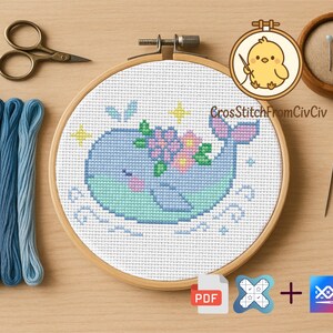 K&ouml;nnte beinhalten: Ein Kreuzstichprojekt mit einem blauen Wal mit floralen Akzenten, pr&auml;sentiert in einem Holzreifen. Das Bild zeigt blaue Stickgarne, eine Schere, Stecknadeln, eine Nadel und das Logo "CrossStitchFromCivCiv".