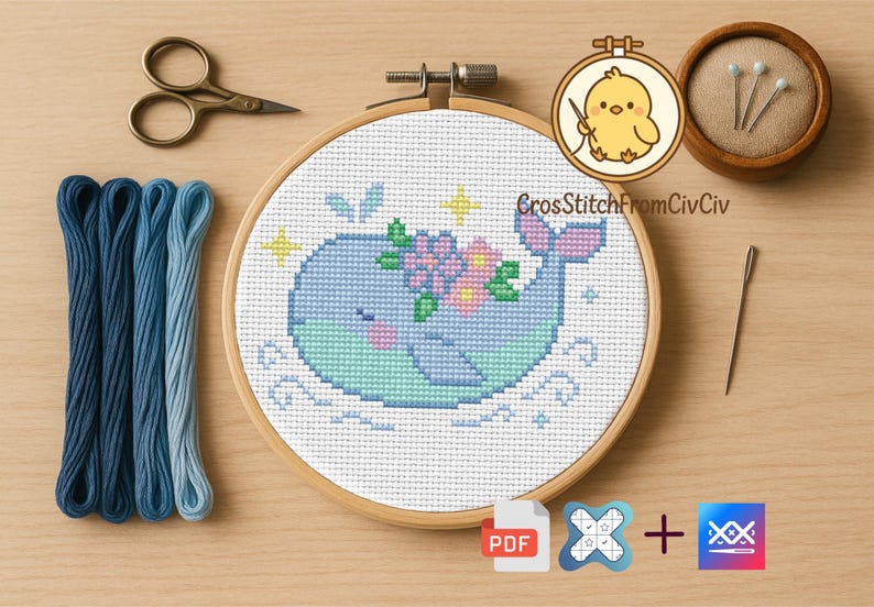 K&ouml;nnte beinhalten: Ein Kreuzstichprojekt mit einem blauen Wal mit floralen Akzenten, pr&auml;sentiert in einem Holzreifen. Das Bild zeigt blaue Stickgarne, eine Schere, Stecknadeln, eine Nadel und das Logo "CrossStitchFromCivCiv".