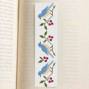 May include: A white bookmark with a cross-stitch design featuring three bluebirds perched on branches with red berries. The bookmark is rectangular and is placed in an open book.
