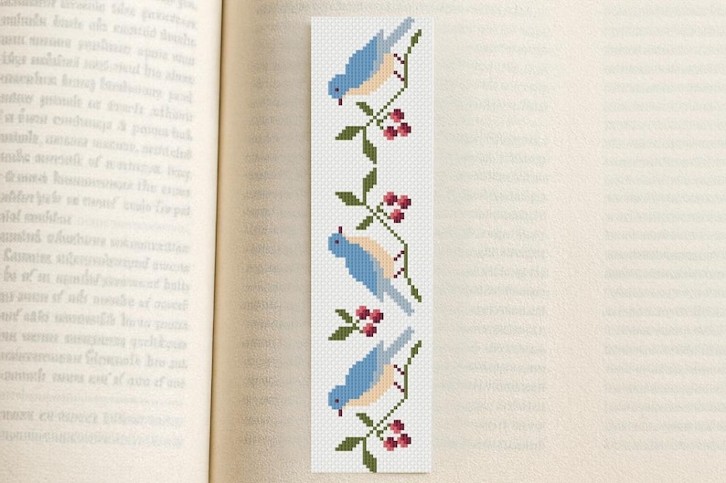 May include: A white bookmark with a cross-stitch design featuring three bluebirds perched on branches with red berries. The bookmark is rectangular and is placed in an open book.