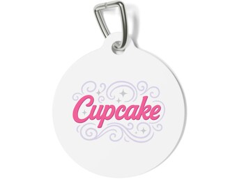 May include: A white, round pet tag with a silver-colored metal loop. The tag features the word "Cupcake" in pink script, surrounded by decorative swirls and stars in a light purple color.