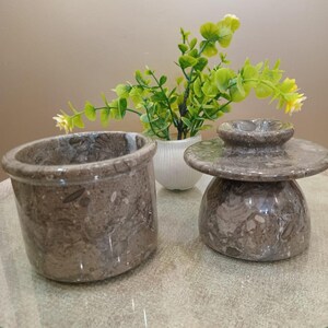 May include: Two pieces of gray marble kitchenware. One is a cylindrical container, and the other is a covered butter dish. The marble has a speckled pattern. A small white vase with green and yellow plants is in the background.
