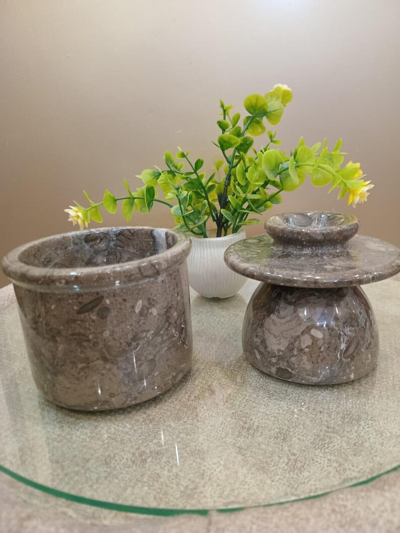 May include: Two pieces of gray marble kitchenware. One is a cylindrical container, and the other is a covered butter dish. The marble has a speckled pattern. A small white vase with green and yellow plants is in the background.