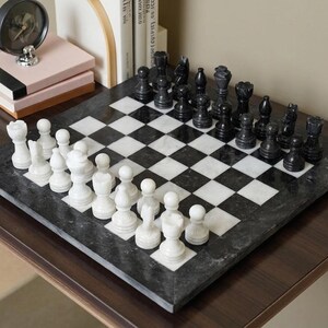 Pu&ograve; includere: Una scacchiera quadrata in marmo lucido bianco e nero con motivo a scacchi. La scacchiera presenta un set completo di pezzi degli scacchi in pietra intagliata nei colori bianco e nero, disposti nelle loro posizioni iniziali sulla superficie.