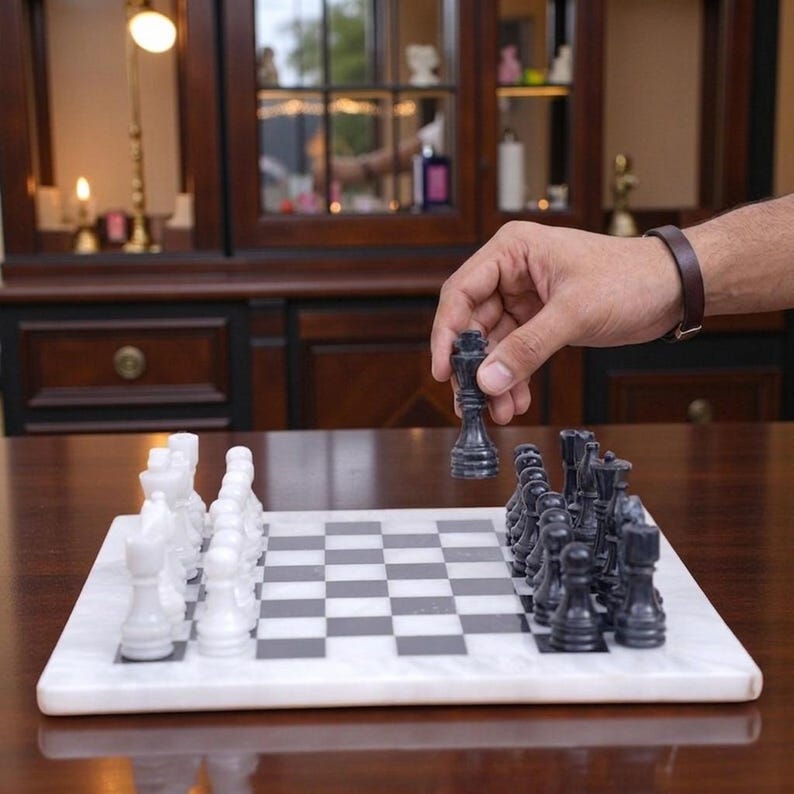 Pu&ograve; includere: Un set di scacchi in marmo con una scacchiera quadrata a caselle bianche e nere alternate. Il set include una collezione completa di pezzi in pietra lucida bianchi e neri, con un re nero tenuto sospeso sopra la scacchiera.