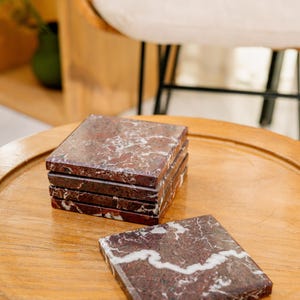 Pu&ograve; includere: Un set di cinque sottobicchieri quadrati in marmo rosso scuro lucido con venature bianche prominenti. Quattro sottobicchieri sono impilati in una pila ordinata, mentre un singolo sottobicchiere &egrave; appoggiato in piano davanti, mostrando la pietra.