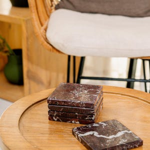 Pu&ograve; includere: Un set di cinque sottobicchieri quadrati in marmo rosso scuro con venature bianche. Quattro sottobicchieri sono impilati verticalmente, mentre uno &egrave; appoggiato in piano accanto alla pila, mostrando la superficie lucida e i motivi naturali.