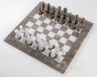 Può includere: Un set di scacchi quadrato con una scacchiera a riquadri alternati in pietra bianca e pietra marrone screziata. Il set include sedici pezzi degli scacchi in pietra bianca e sedici in pietra marrone, intagliati con dettagli arrotondati.