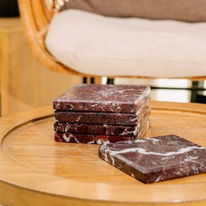 Pu&ograve; includere: Un set di cinque sottobicchieri quadrati in marmo rosso scuro con venature bianche prominenti. Quattro sottobicchieri sono impilati verticalmente in una colonna, mentre il quinto &egrave; appoggiato in piano accanto alla pila, mostrando la texture della pietra lucida.
