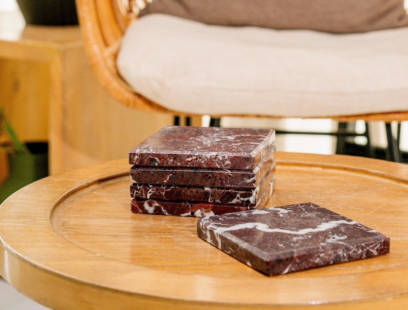 Pu&ograve; includere: Un set di cinque sottobicchieri quadrati in marmo rosso scuro con venature bianche prominenti. Quattro sottobicchieri sono impilati verticalmente in una colonna, mentre il quinto &egrave; appoggiato in piano accanto alla pila, mostrando la texture della pietra lucida.