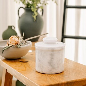 May include: A white marble butter dish with a lid sits on a wooden surface. The dish has a cylindrical shape with a small, round knob on top. The marble has a subtle, swirling pattern. Other items in the background include vases and a bowl with a flower.
