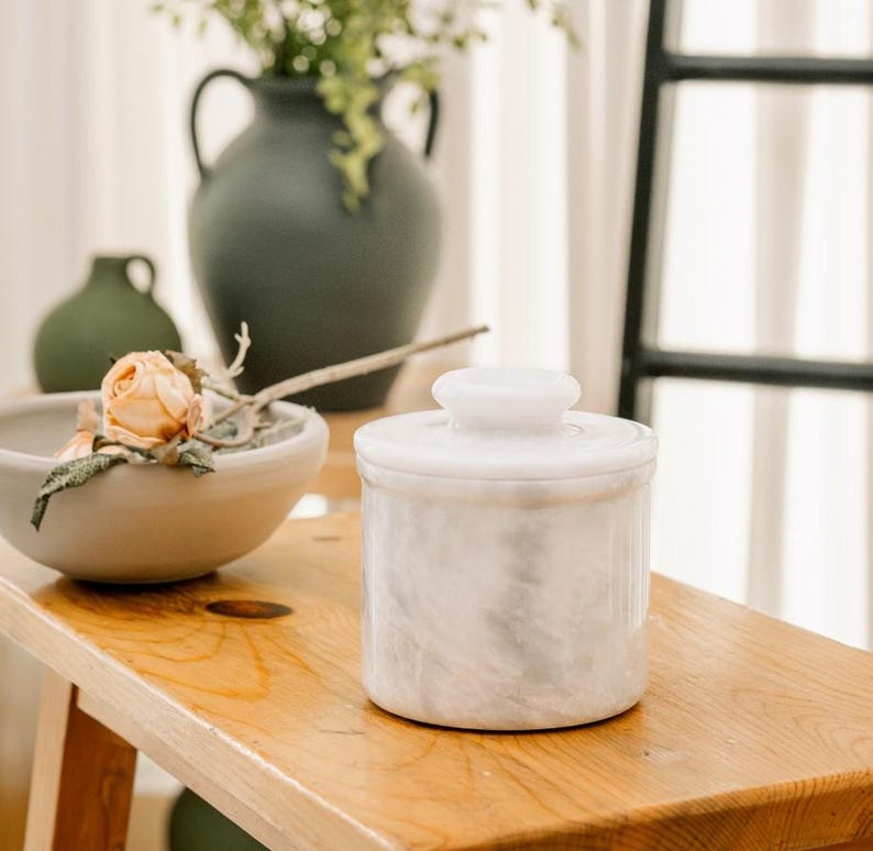 May include: A white marble butter dish with a lid sits on a wooden surface. The dish has a cylindrical shape with a small, round knob on top. The marble has a subtle, swirling pattern. Other items in the background include vases and a bowl with a flower.