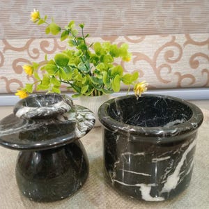 May include: Two black marble containers with white veining. One is a small, lidded jar, and the other is a cylindrical pot. The containers are on a light-colored surface, with a small plant in the background.